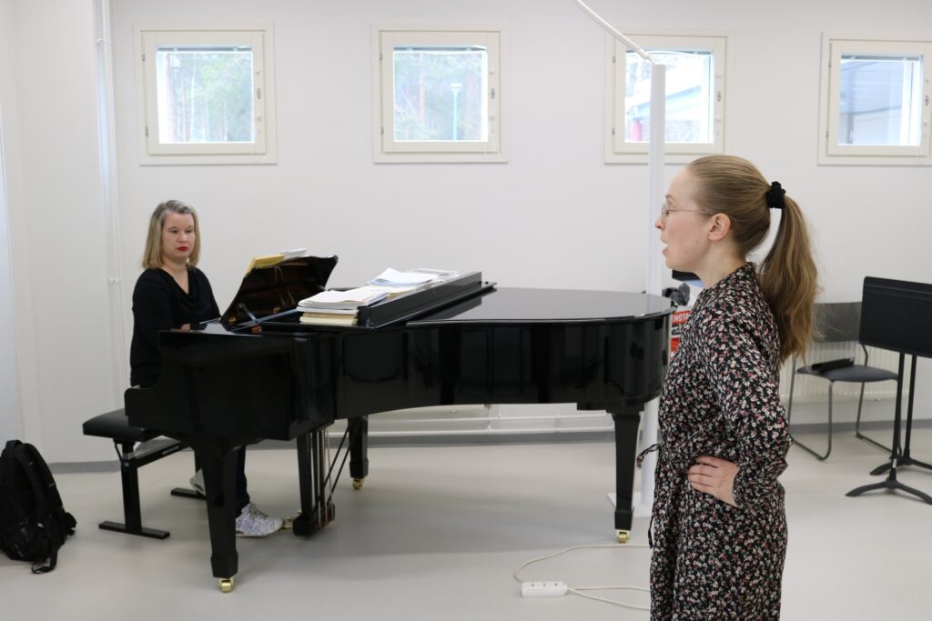 Vasemmalla pianoa soittava henkilö, oikealla etualalla laulaja sivuttain kameraan nähden.