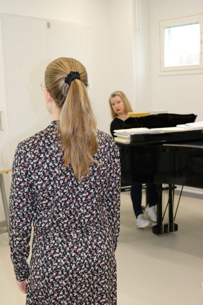 Etualalla laulaja selin kameraan, taustalla pianoa soittava henkilö.