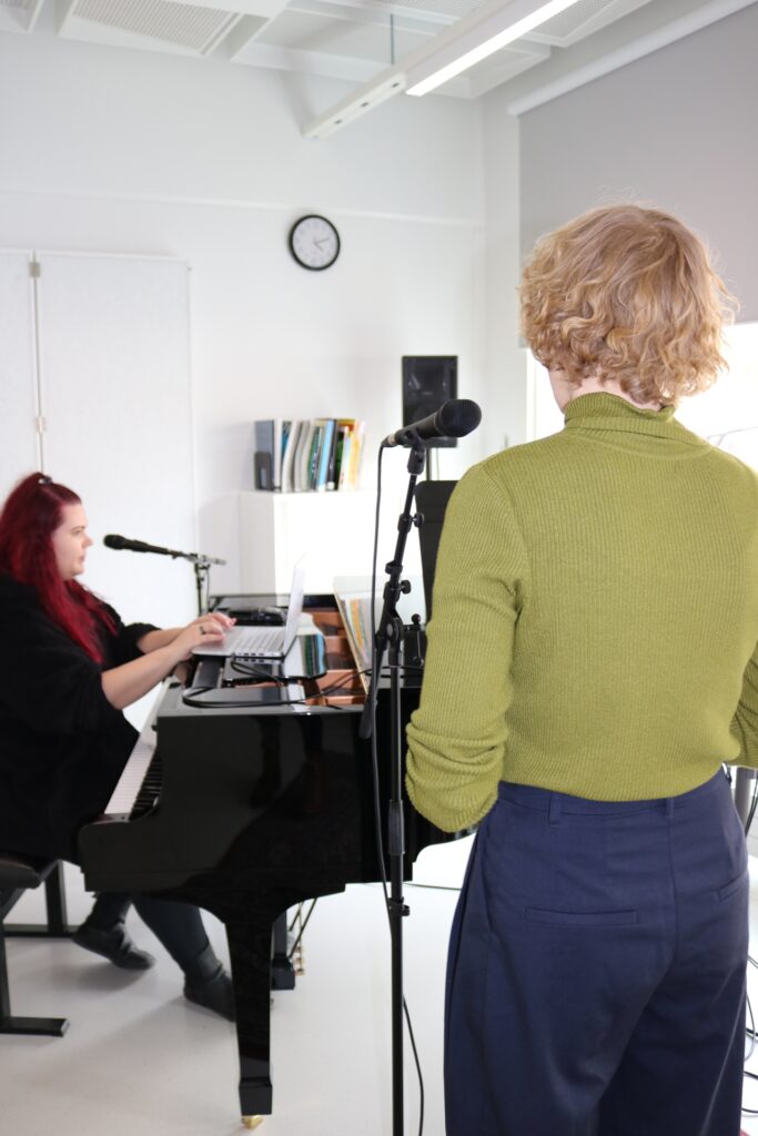 Etualalla laulutunnilla oleva laulaja selkä kameraa kohden, taustalla pianon ääressä toinen henkilö.