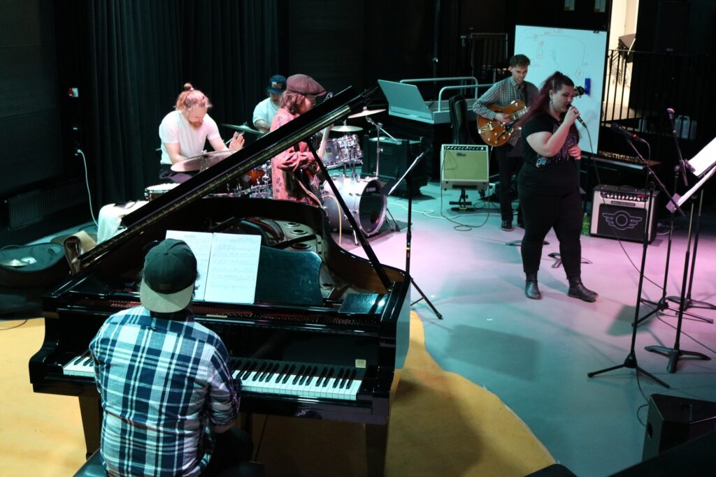 Esiintymistä harjoitteleva yhtye, johon kuuluu etualalla pianisti ja taaempana kaksi rumpalia, kaksi kitaristia sekä laulaja.