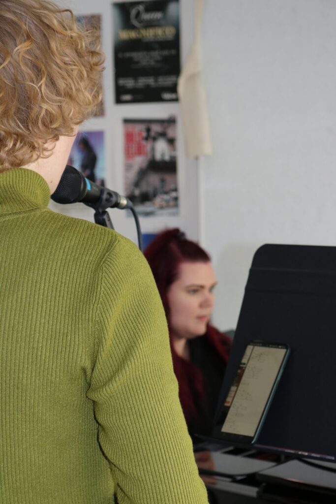 Etualalla laulutunnilla oleva laulaja selkä kameraa kohti, taustalla henkilö soittamassa pianoa.