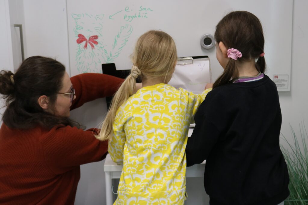 Selkä kameraan opettaja ja kaksi lasta oppitunnilla.